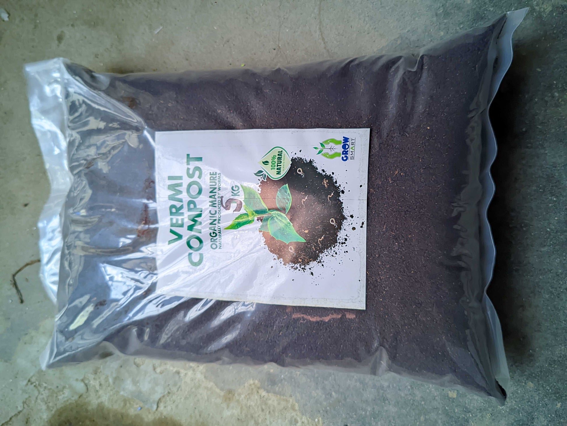 A large clear plastic bag filled with dark brown vermi compost manure, featuring a white label with green and black text and a small green leaf logo, placed on a concrete floor.